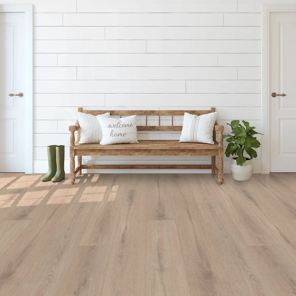 A wooden bench with white pillows and a "welcome home" cushion sits between two doors, highlighting the craftsmanship of a leading flooring company in Mississippi. A pair of green boots and a potted plant rest on the exquisite floor.