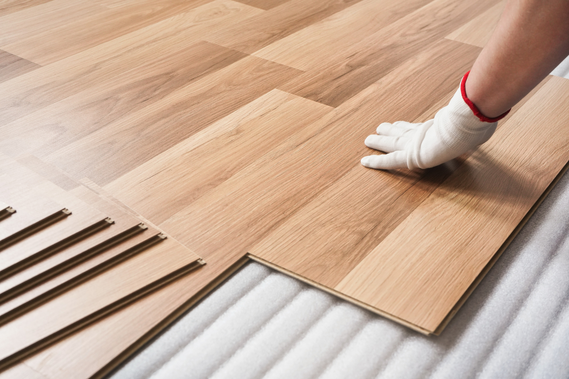 A person wearing a white glove is carefully installing interlocking wooden laminate flooring, provided by a leading flooring supplier, with several boards laid out and aligned on a white foam underlayment.