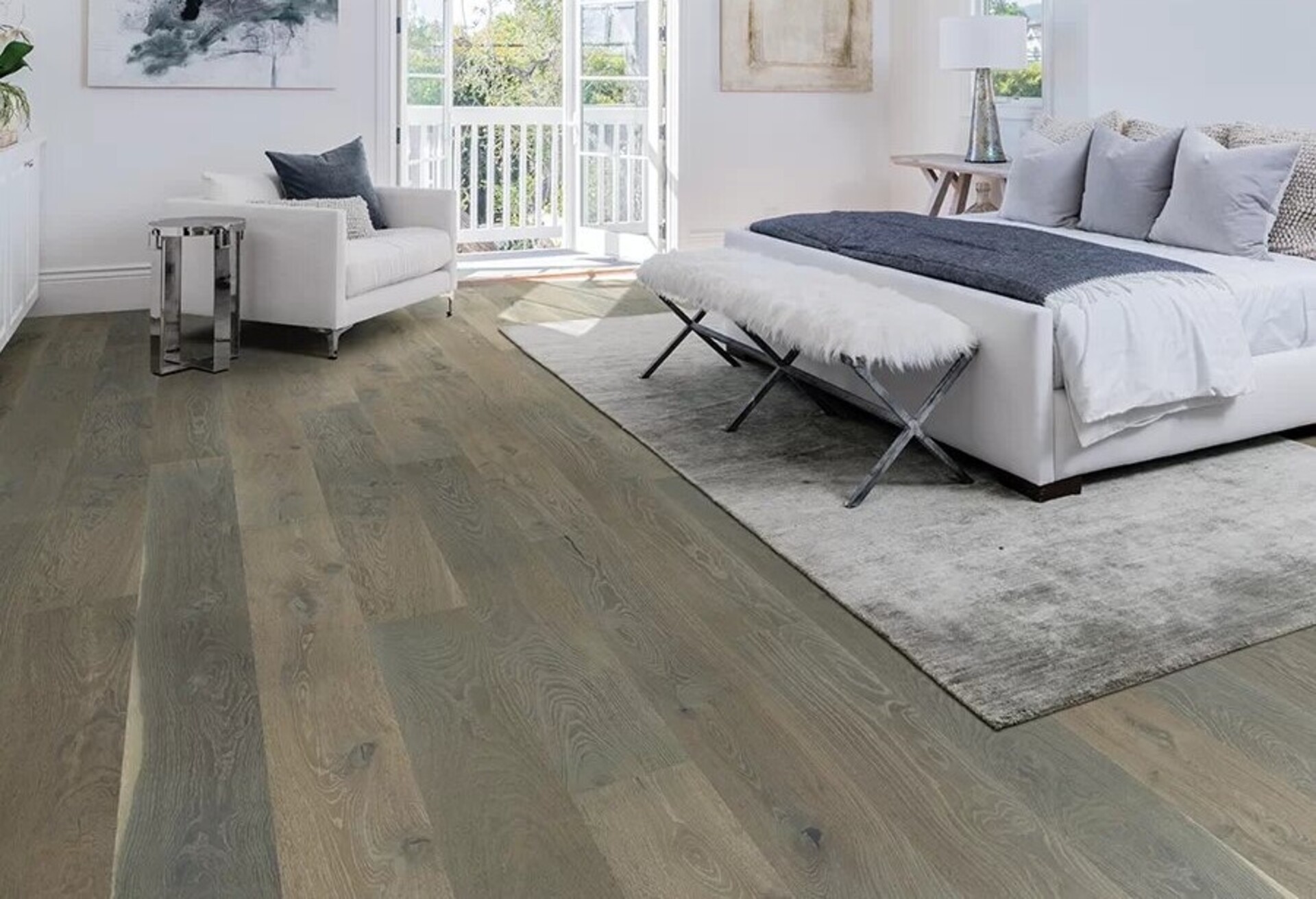 A bright bedroom featuring a large bed with gray bedding and a white armchair on elegant hardwood floors. Courtesy of a renowned flooring supplier, the space opens to a balcony with lush greenery outside.