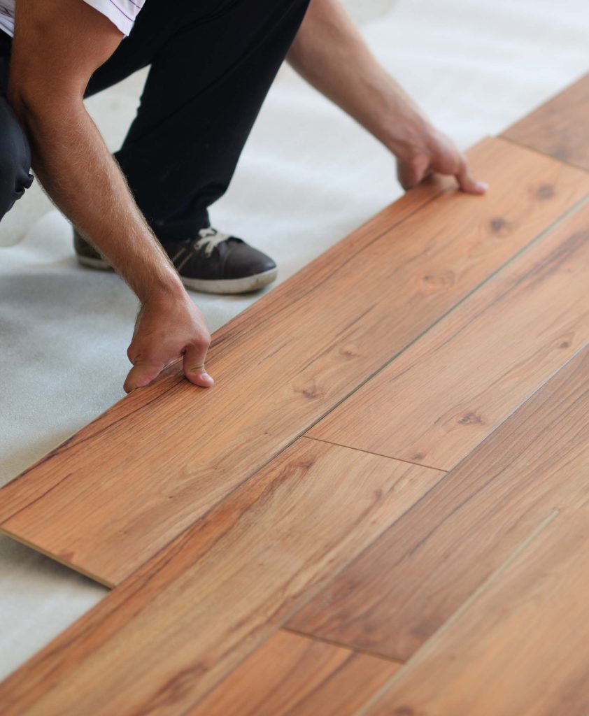 A person meticulously installing wooden laminate flooring, their skilled hands placing a plank with precision on the partially finished surface. This scene captures the craftsmanship of a reputable flooring installation company serving Mississippi and Louisiana.