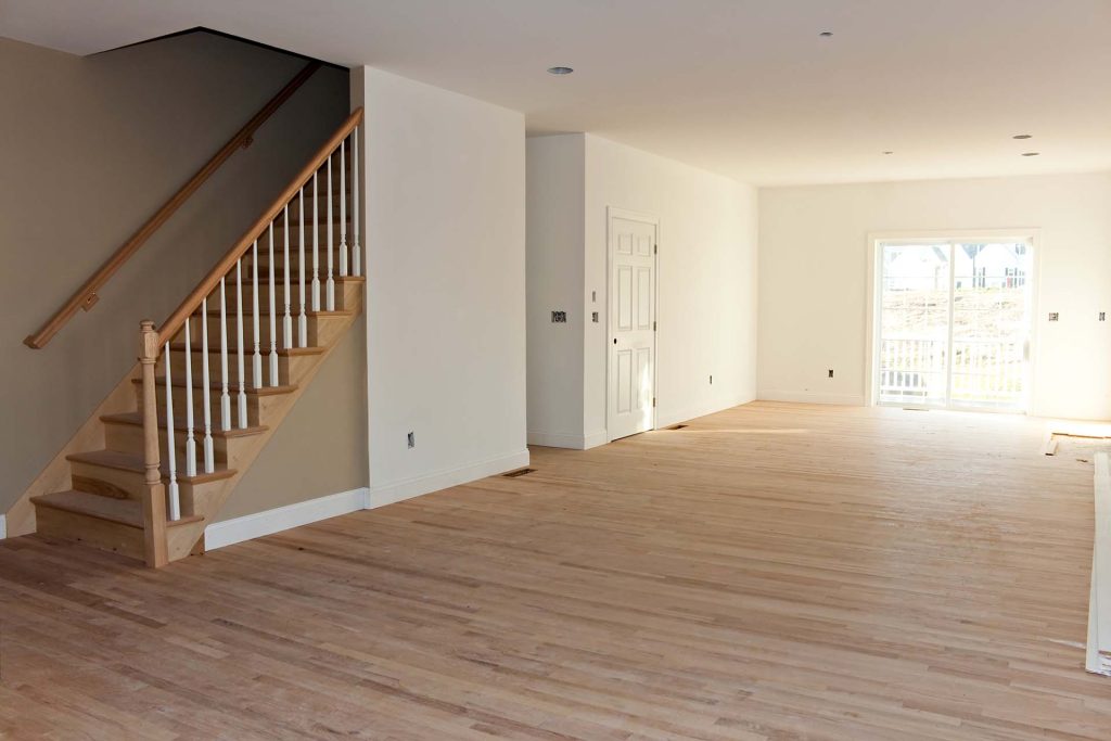 An empty room with light wood floors crafted by a renowned flooring company, a staircase with wooden railing on the left, pristine white walls, and a sliding glass door leading outside.