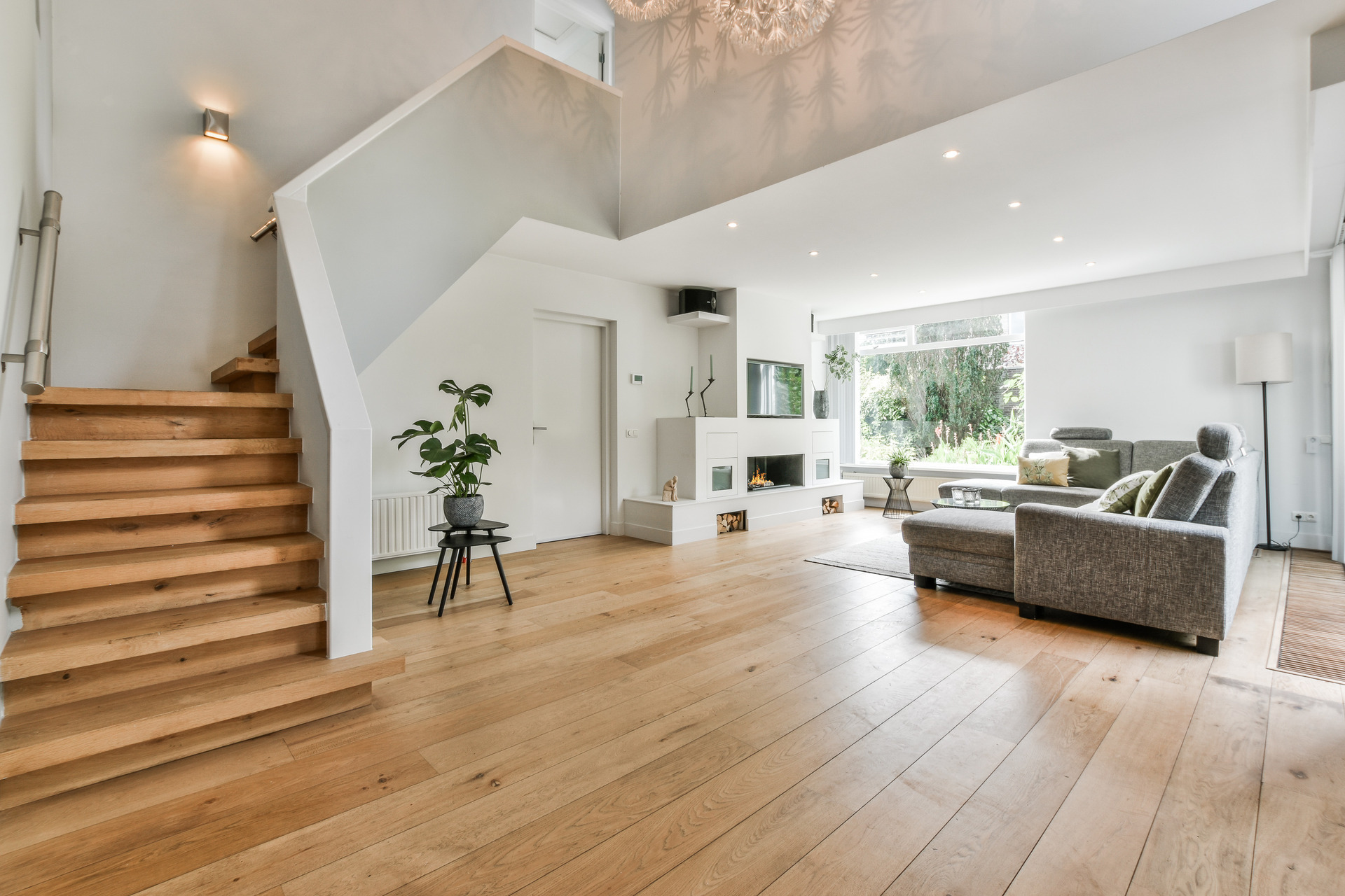 Modern living room with wooden flooring crafted by a top flooring company, featuring a gray sectional sofa, a small plant on a stand, and a staircase leading to an upper level.