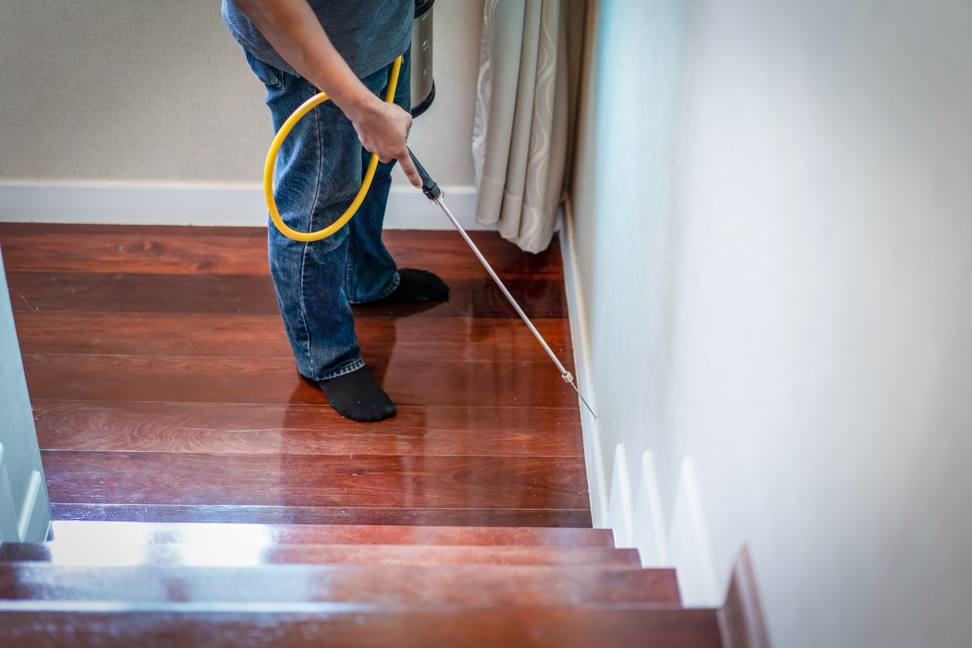 A flooring company expert uses a spray wand to apply a substance along the base of a wall on a wooden staircase.