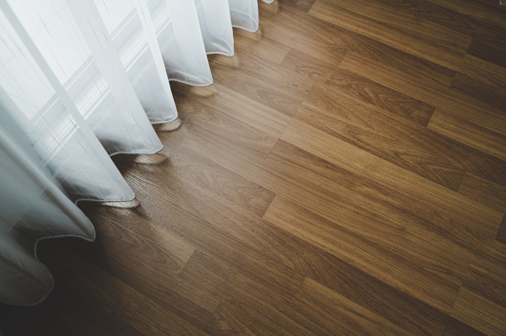 Sunlight streams through sheer white curtains, illuminating the light brown wooden floor crafted by a top-tier flooring company.