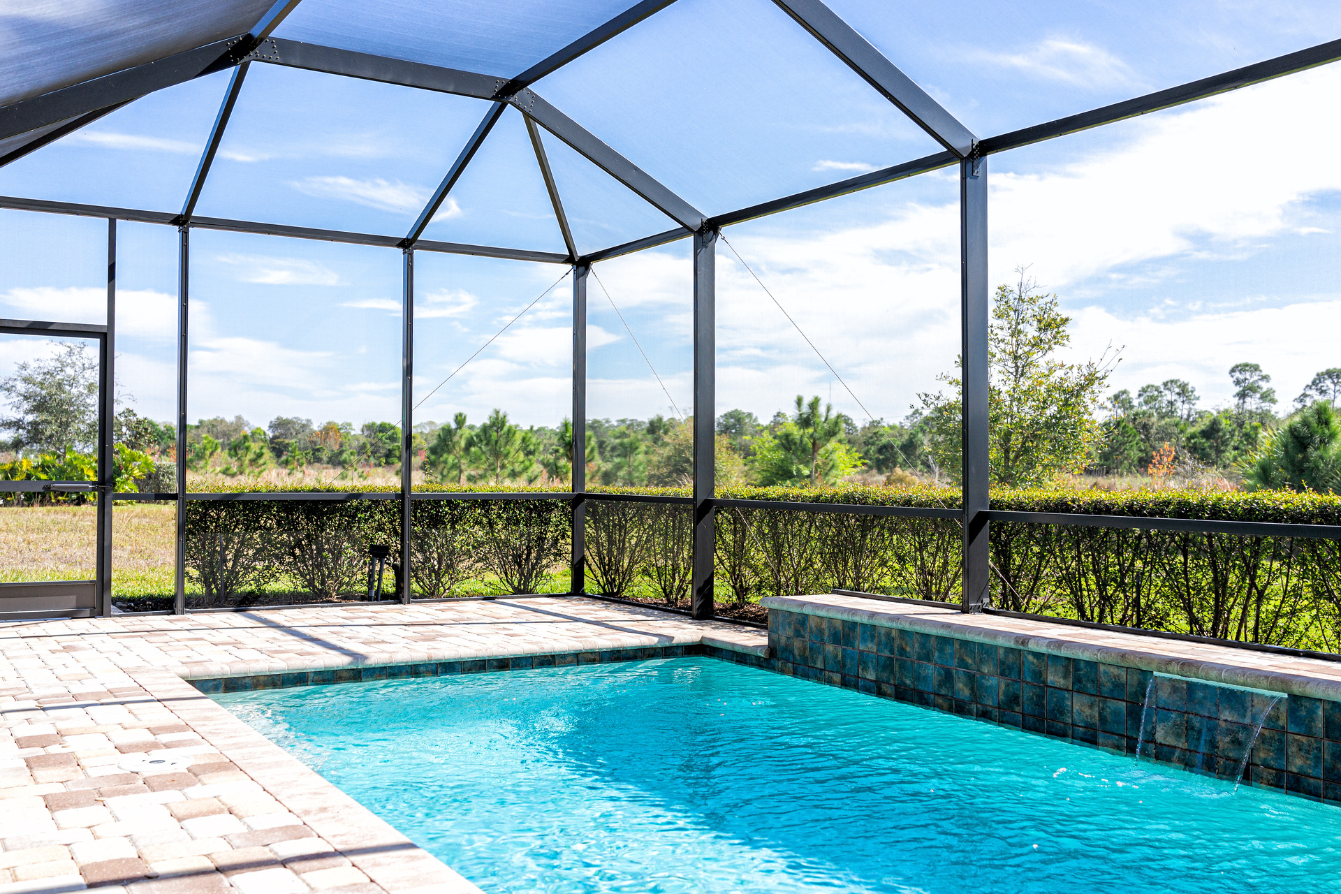 A screened-in outdoor pool with a tile deck supplied by a trusted flooring company in Mississippi overlooks a natural landscape with trees and a clear blue sky.