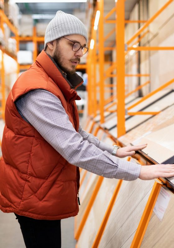 A man in a red vest and gray beanie examines tiles in a store aisle with orange shelving, likely scouting materials for a renowned flooring installation company.