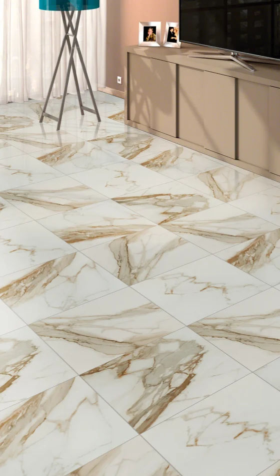 A stunning living room features beige marble tile flooring expertly crafted by a premier flooring installation company. With a beige TV stand, flat-screen TV, and white couch, large windows adorned with sheer curtains provide a serene backdrop.